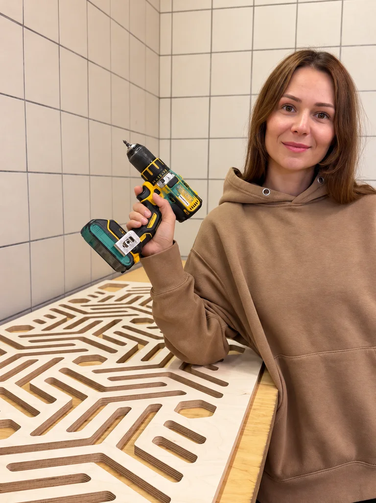 Alisa Shutova holding a cordless drill next to a CNC-cut plywood panel in the workshop